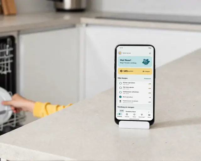 Een kinderhand zet een bord in de vaatwasser. Op het aanrecht staat een telefoon met AiFamQuest: Hoi Noor, nog 3 klusjes vandaag, 185 punten.